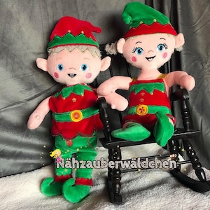 May include: Two plush elf dolls wearing red and green elf costumes with pointy hats. The dolls are sitting on a wooden rocking chair. The chair is painted black and has a white sign that says "Nähzauberwäldchen".