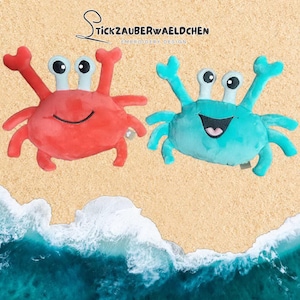 Pode incluir: Dois brinquedos de pelúcia em forma de caranguejo, um vermelho e outro azul-turquesa, com rostos sorridentes e olhos arregalados. Os caranguejos estão em um fundo de praia arenosa com ondas do oceano. O texto "Stickzauberwaeldchen Embroidery Design" está no topo.