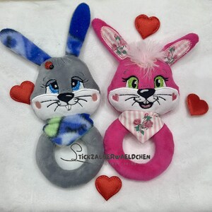 Puede incluir: Dos juguetes de peluche con forma de conejo, uno gris con una bufanda azul y otro rosa con una bufanda rosa y blanca. Ambos tienen caras bordadas y est&aacute;n rodeados de corazones de fieltro rojo.