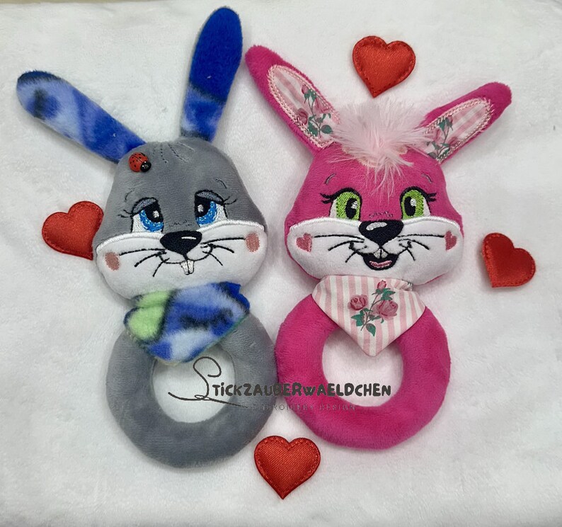 Puede incluir: Dos juguetes de peluche con forma de conejo, uno gris con una bufanda azul y otro rosa con una bufanda rosa y blanca. Ambos tienen caras bordadas y est&aacute;n rodeados de corazones de fieltro rojo.