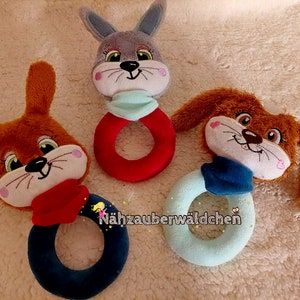 Puede incluir: Tres juguetes de anillo de animales de peluche. Un conejito marr&oacute;n con una bufanda roja, un conejito gris con una bufanda azul y un perro marr&oacute;n con una bufanda azul. Cada juguete tiene un anillo unido a la parte inferior.