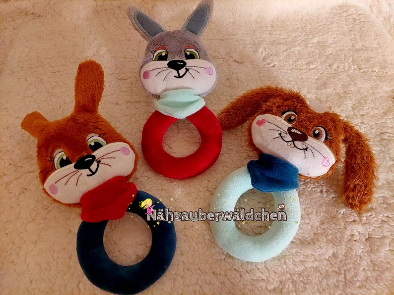 Puede incluir: Tres juguetes de anillo de animales de peluche. Un conejito marr&oacute;n con una bufanda roja, un conejito gris con una bufanda azul y un perro marr&oacute;n con una bufanda azul. Cada juguete tiene un anillo unido a la parte inferior.