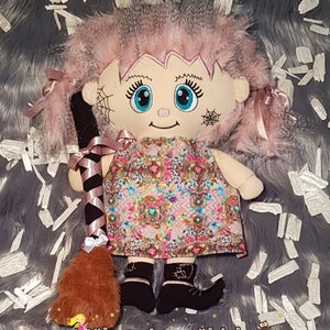 Könnte beinhalten: Handgemachte Puppe mit rosa Haaren, blauen Augen und einem Blumenkleid. Die Puppe hat ein gesticktes Gesicht mit Spinnennetz-Design und hält einen schwarz-rosa Besen. Die schwarzen Stiefel der Puppe haben ein Herz-Design. Der Text "Nähzauberwäldchen" steht unten.