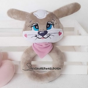 Puede incluir: Un juguete de anillo de peluche gris y blanco con ojos bordados, una nariz rosa y una bandana rosa. El juguete est&aacute; dise&ntilde;ado para parecer un perro y est&aacute; etiquetado como "Stickzauberwaeldchen Embroidery Design."