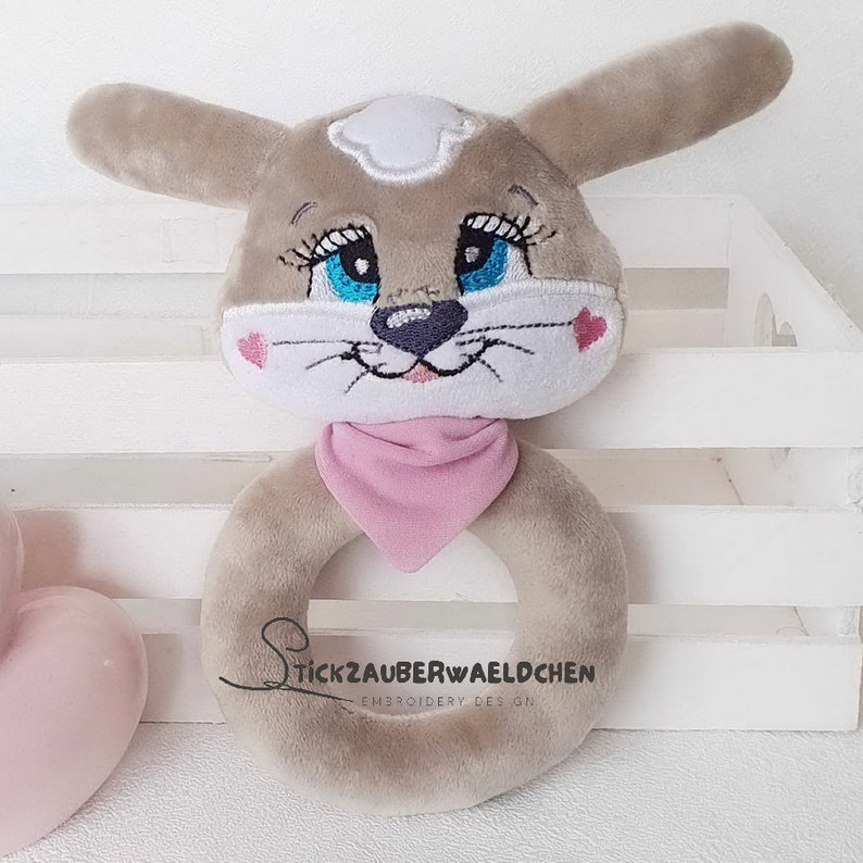 Puede incluir: Un juguete de anillo de peluche gris y blanco con ojos bordados, una nariz rosa y una bandana rosa. El juguete est&aacute; dise&ntilde;ado para parecer un perro y est&aacute; etiquetado como "Stickzauberwaeldchen Embroidery Design."