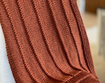 May include: A knitted throw blanket in a warm rust orange color. The blanket has a ribbed texture and is draped over a chair.