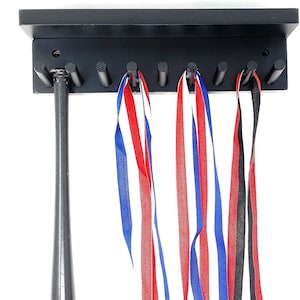 May include: Black wooden wall-mounted medal display with a shelf and hooks. The display has a baseball bat hanging from one hook and four ribbons hanging from other hooks. The ribbons are red, white, and blue.