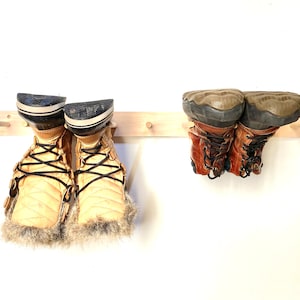 Puede incluir: Un zapatero de madera con dos pares de botas colgadas. El zapatero está montado en una pared blanca. Un par de botas es de cuero marrón con cordones negros, y el otro par es de color beige con cordones negros y ribete de piel.