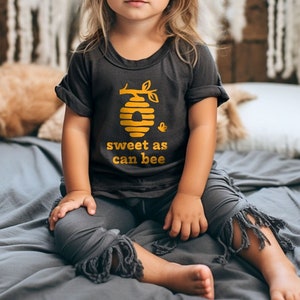 Little girl wearing heather dark grey short sleeve toddler crew neck tee with mustard yellow sweet as can bee text with flying bumble bee outisde beehive hanging off branch print, sitting on bed posing in front of boho decor