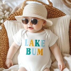 Baby wearing white short sleeve baby bodysuit with alternating light blue, mustard yellow and teal blue LAKE BUM text print on front, laying in wicker basket with blankets and pillow, wearing a sun hat and sunglasses