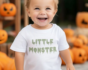 May include: A young child wearing a white t-shirt with the text "LITTLE MONSTER" in green dripping letters. The child is smiling and standing in front of a stack of orange pumpkins.
