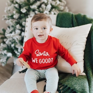 Cute little toddler boy wearing red long sleeve crew neck sweatshirt with white Santa, WE GOOD? graphic print, sitting on chair in front of Christmas tree holding a cookie