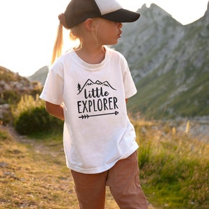 Little girl outside in nature with steep mountains in the background, wearing white short sleeve toddler crew neck tee with printed black LITTLE EXPLORER hand drawn rocky mountain range, pine tree and arrow design