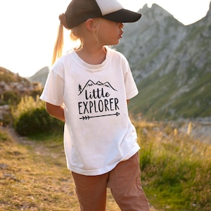 Little girl outside in nature with steep mountains in the background, wearing white short sleeve toddler crew neck tee with printed black LITTLE EXPLORER hand drawn rocky mountain range, pine tree and arrow design