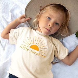 Little girl laying down in grass wearing a hat and natural cream colored short sleeve kids crew neck shirt with mustard yellow little sunshine text print with boho sun rays design