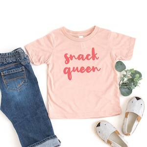 Heather peach short sleeve unisex toddler crew neck tshirt with pink cursive snack queen text design, laying flat on white background with jeans, greenery and cute shoes