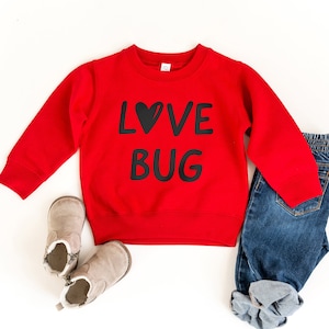 May include: Red sweatshirt with black text that says "LOVE BUG" with a heart replacing the "O". The sweatshirt is paired with blue jeans and brown boots.