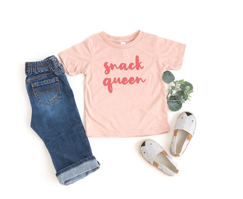 Heather peach short sleeve unisex toddler crew neck tshirt with pink cursive snack queen text design, laying flat on white background with jeans, greenery and cute shoes