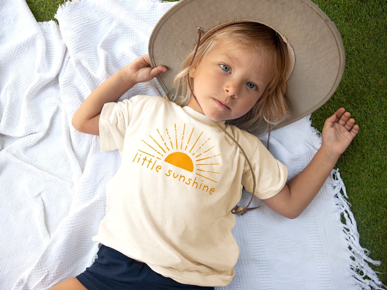 Little girl laying down in grass wearing a hat and natural cream colored short sleeve kids crew neck shirt with mustard yellow little sunshine text print with boho sun rays design