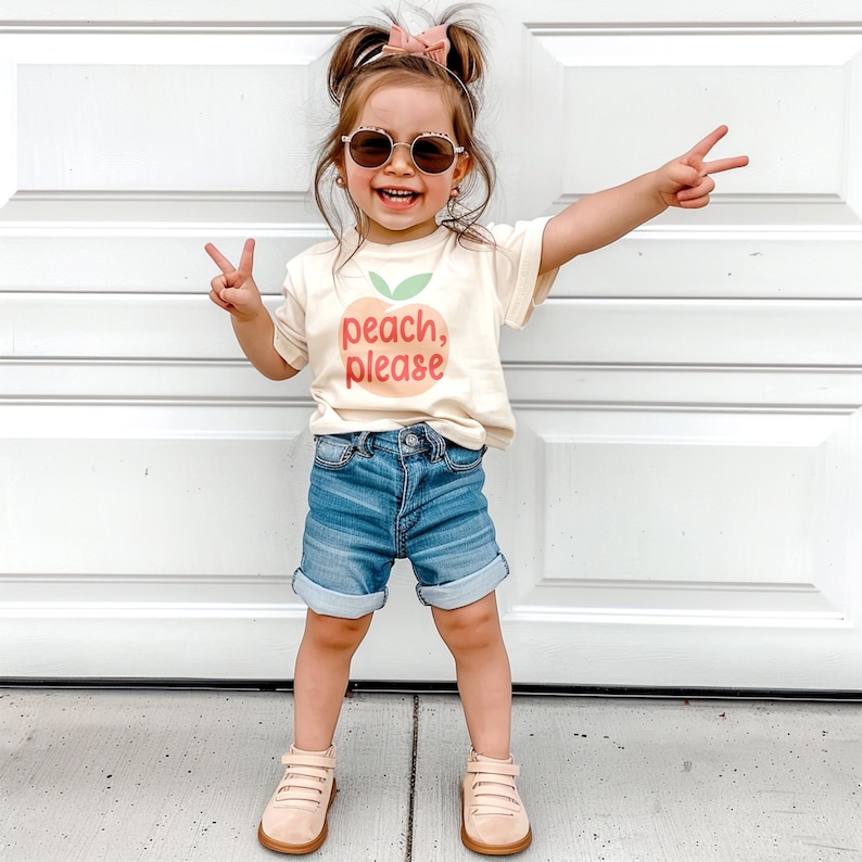 Cute toddler girl wearing short sleeve natural white tshirt with graphic print of a light peach and light green peach fruit with pink peach, please text print in front, standing outside with a pink bow and sunglasses holding up peace signs