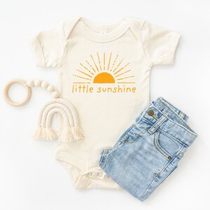 Natural white short sleeve infant bodysuit with mustard yellow little sunshine text with boho sun rays design printed on front, laying flat on white background with jeans and woven rainbow teething toy