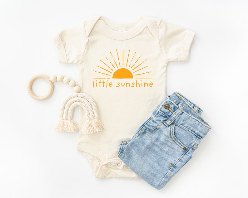 Natural white short sleeve infant bodysuit with mustard yellow little sunshine text with boho sun rays design printed on front, laying flat on white background with jeans and woven rainbow teething toy