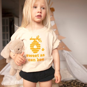 Cute little girl wearing natural white short sleeve toddler crew neck tee with mustard yellow sweet as can bee text with flying bumble bee outisde beehive hanging off branch print, standing in front of play teepee holding bunny stuffed animal