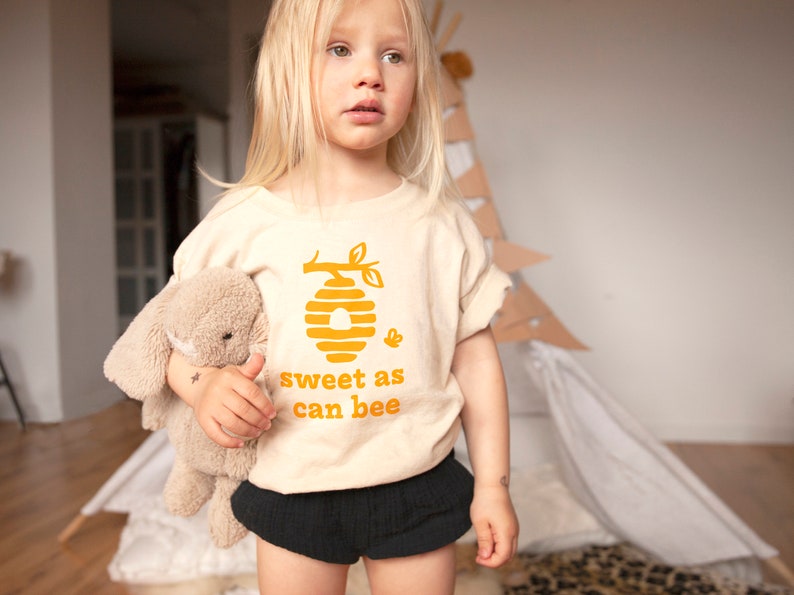 Cute little girl wearing natural white short sleeve toddler crew neck tee with mustard yellow sweet as can bee text with flying bumble bee outisde beehive hanging off branch print, standing in front of play teepee holding bunny stuffed animal