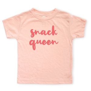 Heather peach short sleeve unisex toddler crew neck tshirt with pink cursive snack queen text design, laying flat on white background