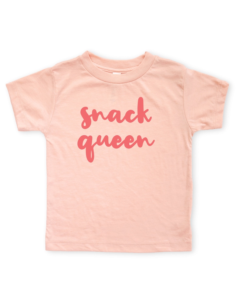 Heather peach short sleeve unisex toddler crew neck tshirt with pink cursive snack queen text design, laying flat on white background