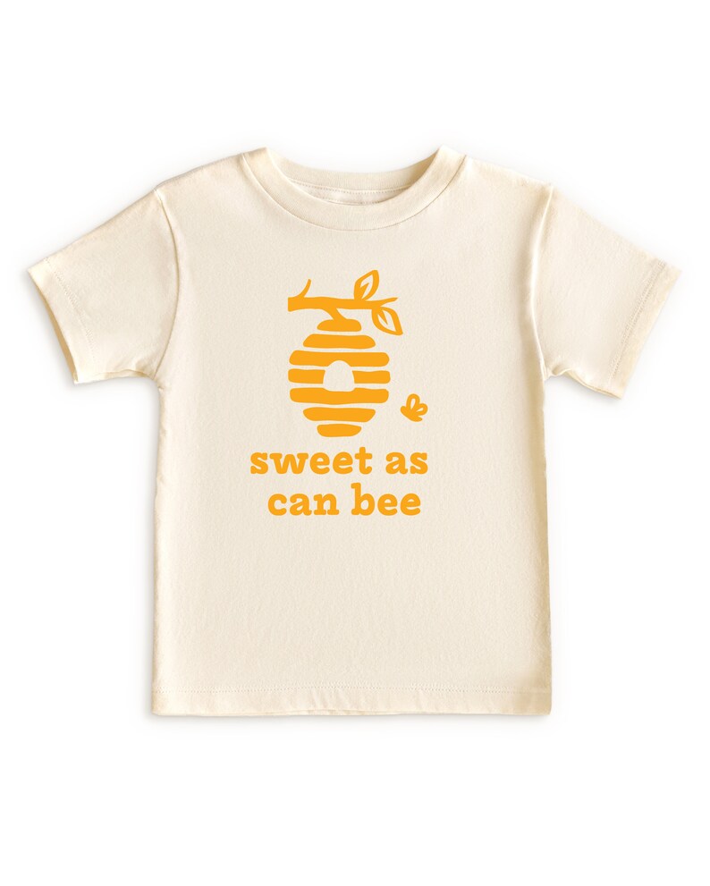 Natural white short sleeve toddler crew neck tee with mustard yellow sweet as can bee text with flying bumble bee outisde beehive hanging off branch print, laying flat on white background