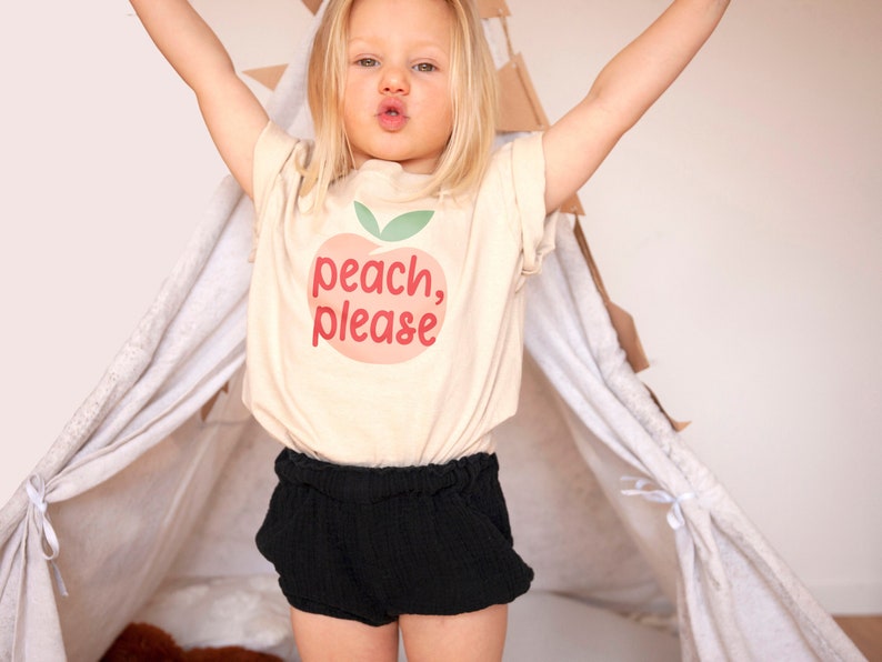 Cute toddler girl wearing short sleeve natural white tshirt with graphic print of a light peach and light green peach fruit with pink peach, please text print in front, standing in front of teepee with hands in the air
