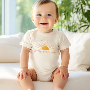 Cute baby wearing natural cream colored short sleeve infant bodysuit with mustard yellow little sunshine text with boho sun rays design printed on front, sitting on couch in front of window with greenery plants