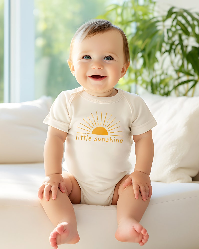 Cute baby wearing natural cream colored short sleeve infant bodysuit with mustard yellow little sunshine text with boho sun rays design printed on front, sitting on couch in front of window with greenery plants