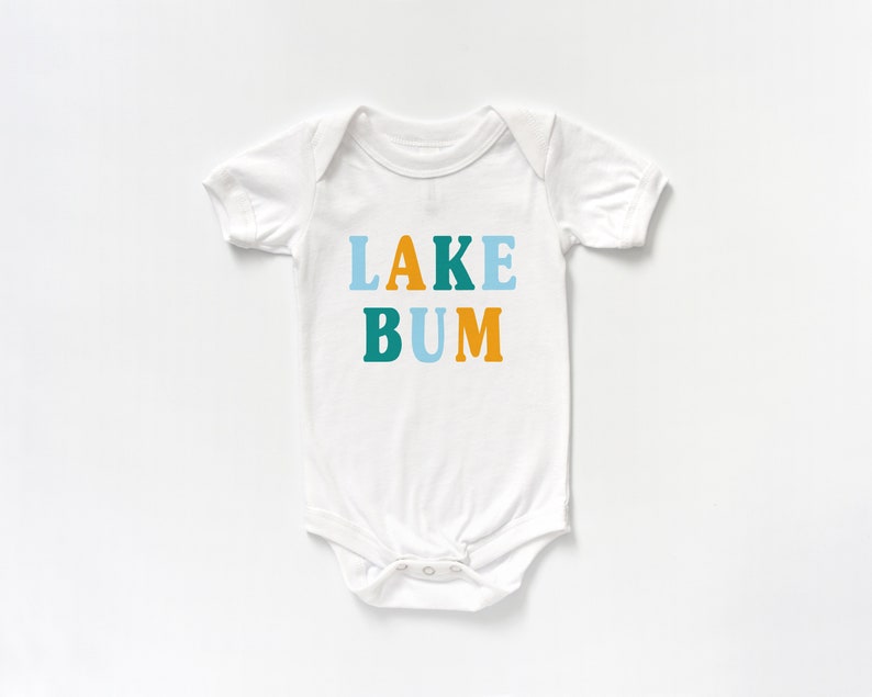 White short sleeve baby bodysuit with alternating light blue, mustard yellow and teal blue LAKE BUM text print on front, laying flat on white background