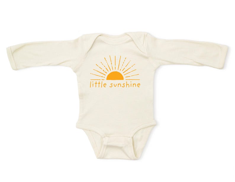 K&ouml;nnte beinhalten: Ein wei&szlig;er lang&auml;rmeliger Baby-Strampler mit einem orangefarbenen Sonnenmotiv und dem Text "little sunshine".