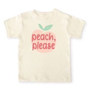 Short sleeve natural white toddler tshirt with graphic print of a light peach and light green peach fruit with pink peach, please text print in front, laying flat on white background