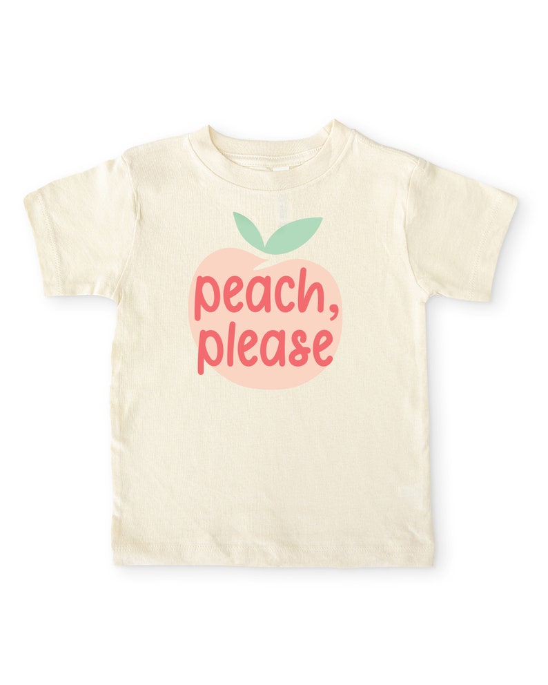 Short sleeve natural white toddler tshirt with graphic print of a light peach and light green peach fruit with pink peach, please text print in front, laying flat on white background