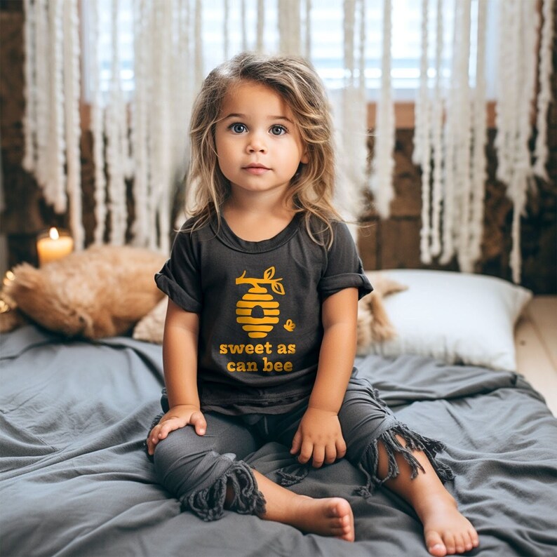 Little girl wearing heather dark grey short sleeve toddler crew neck tee with mustard yellow sweet as can bee text with flying bumble bee outisde beehive hanging off branch print, sitting on bed posing in front of boho decor