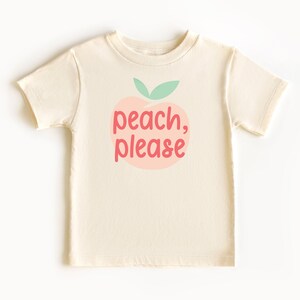 Short sleeve natural white toddler tshirt with graphic print of a light peach and light green peach fruit with pink peach, please text print in front, laying flat on white background
