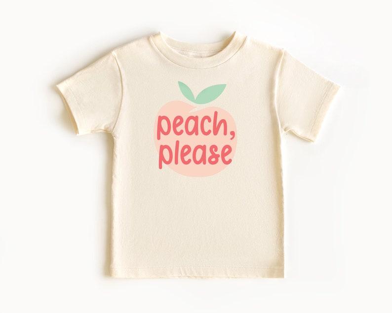 Short sleeve natural white toddler tshirt with graphic print of a light peach and light green peach fruit with pink peach, please text print in front, laying flat on white background