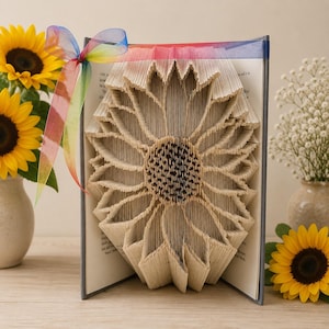 May include: A book with pages folded to form a three-dimensional sunflower shape. The book is adorned with a translucent rainbow-colored ribbon tied in a bow at the top corner. The paper pages are cream-colored with visible black printed text.