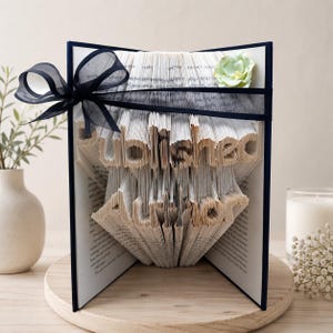 May include: A book sculpture featuring the words Published Author created by folding individual pages. The book is bound in dark blue covers and decorated with a sheer black ribbon tied in a bow and a single pale green fabric flower on the top right.
