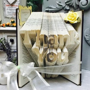 May include: A book with the words "Thank You" folded into the pages. The book is decorated with a white ribbon and a yellow flower.