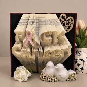 May include: A folded book art sculpture displaying the text FUCK CANCER with a pink ribbon attached to the front. The sculpture is positioned next to a white ceramic pitcher with cow patterns, a white rose, and two white ceramic bird figurines.