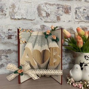 May include: A book art sculpture with the letters "LOVE" folded from the pages. The book is open and the pages are fanned out to create the letters. The sculpture is decorated with small artificial flowers and a lace ribbon. The book is sitting on a table with a white background.