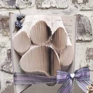 May include: A unique book art piece featuring a paw print design. The pages of the book are folded to create the paw print shape. A lavender ribbon and bow are attached to the book, along with a decorative floral accent.