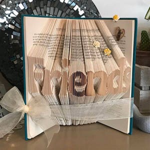 May include: A book art sculpture with the word "Friends" folded into the pages. The book is bound in teal and has a white ribbon tied around it. The sculpture is decorated with small yellow flowers.