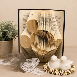May include: A folded book art sculpture featuring the silhouette of a cartoon mouse head, displayed with a black cover. Beside it sits a pair of white ceramic bird figurines nestled in a ring of small pink and green decorative floral beads.