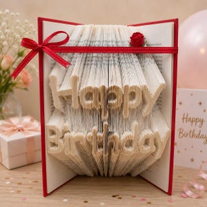 May include: A folded book art sculpture with pages intricately creased to form the words Happy Birthday in a stacked layout. The book features a red hard cover, a red ribbon tied in a bow on the left side, and a small red fabric rose on the top right.